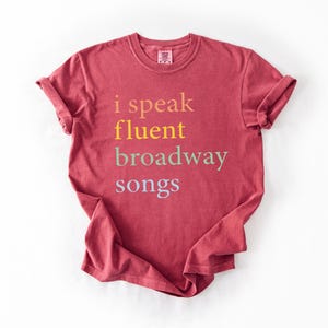 May include: A maroon t-shirt with the text "i speak fluent broadway songs" printed in a rainbow of colors.