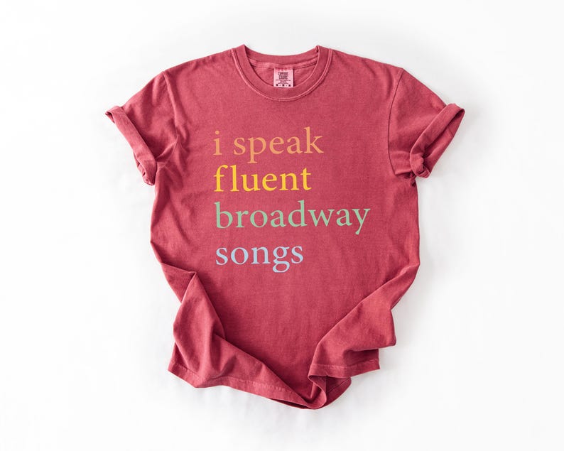 May include: A maroon t-shirt with the text "i speak fluent broadway songs" printed in a rainbow of colors.