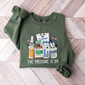 Puede incluir: Sudadera verde con un gr&aacute;fico de cuatro viales de dibujos animados y una bolsa de soluci&oacute;n de dextrosa. El texto "The Pressure Is On" est&aacute; impreso debajo del gr&aacute;fico.