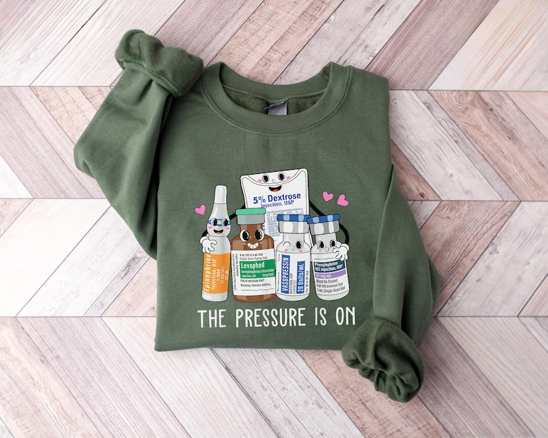 Puede incluir: Sudadera verde con un gr&aacute;fico de cuatro viales de dibujos animados y una bolsa de soluci&oacute;n de dextrosa. El texto "The Pressure Is On" est&aacute; impreso debajo del gr&aacute;fico.