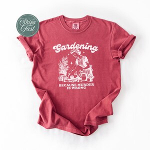 Peut inclure: T-shirt rouge avec l'inscription blanche "Gardening" et "BECAUSE MURDER IS WRONG". Le t-shirt pr&eacute;sente une illustration blanche d'une personne jardinant.