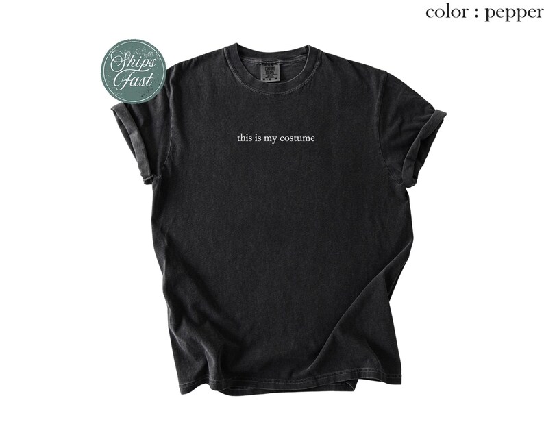 May include: A black, short-sleeved t-shirt with the phrase "this is my costume" printed in white. The shirt has a slightly faded appearance and rolled-up sleeves. The color is pepper.