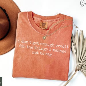 Puede incluir: Una camiseta de color coral con el texto "i don't get enough credit for the things i manage not to say" en blanco. La camiseta tiene cuello redondo y una peque&ntilde;a etiqueta. Un collar dorado con un colgante est&aacute; sobre la camiseta.