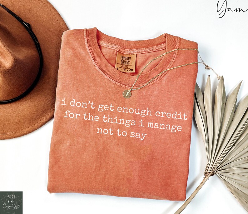 Puede incluir: Una camiseta de color coral con el texto "i don't get enough credit for the things i manage not to say" en blanco. La camiseta tiene cuello redondo y una peque&ntilde;a etiqueta. Un collar dorado con un colgante est&aacute; sobre la camiseta.