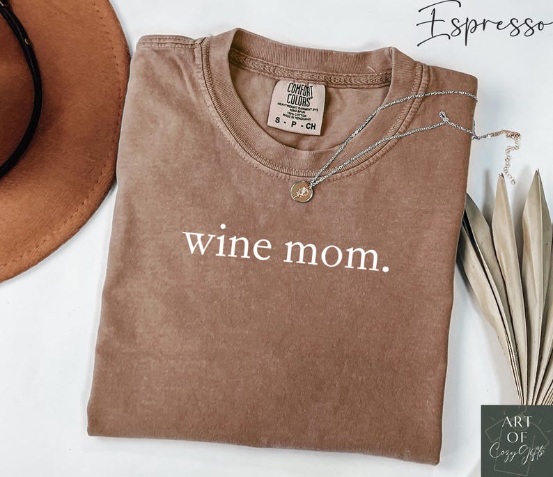 Pu&ograve; includere: Una maglietta marrone con la scritta "wine mom." stampata in bianco. La maglietta &egrave; piegata e appoggiata su una superficie bianca. Si pu&ograve; vedere una collana d'argento con un ciondolo sulla maglietta.