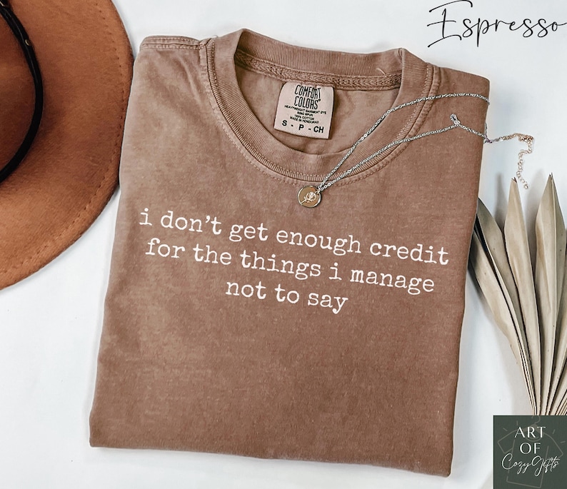 Puede incluir: Camiseta marr&oacute;n con la frase blanca "i don't get enough credit for the things i manage not to say". Un collar plateado con colgante descansa sobre la camiseta. La camiseta tiene una etiqueta Comfort Colors.