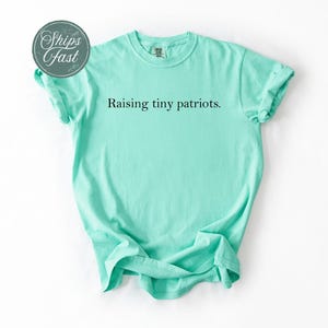 May include: A mint green t-shirt with the phrase "Raising tiny patriots" printed in black. The shirt is folded and laid flat on a white surface. The shirt has short sleeves and a crew neck.