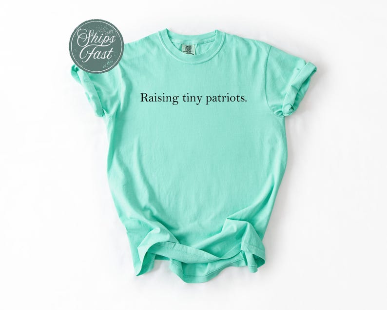 May include: A mint green t-shirt with the phrase "Raising tiny patriots" printed in black. The shirt is folded and laid flat on a white surface. The shirt has short sleeves and a crew neck.