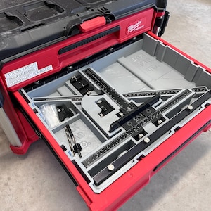 Puede incluir: Una caja de herramientas roja abierta con una tapa superior negra. La caja de herramientas está etiquetada como "Milwaukee Prekout". El interior de la caja de herramientas es gris y contiene una plantilla negra y gris con una regla y una pequeña broca negra. La caja de herramientas también tiene una etiqueta que dice "JAX FAB 20".