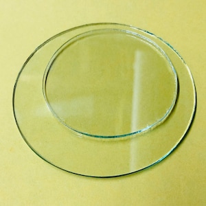May include: Two clear glass plates, one slightly smaller than the other, are stacked on top of each other. The plates are round and have a smooth, polished surface.
