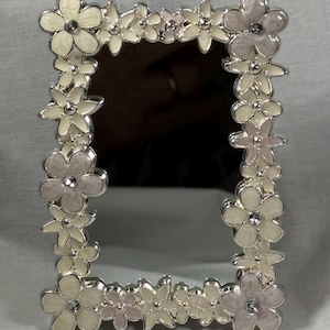 May include: A rectangular mirror with a silver frame adorned with cream and pale pink flower embellishments. Each flower has a small crystal in the center. The frame is made of silver metal.