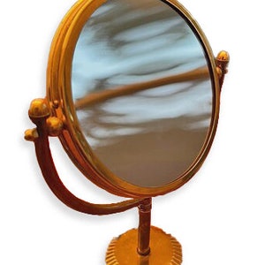 May include: A gold-toned, round, tabletop vanity mirror with a swivel stand. The mirror has a smooth, reflective surface.