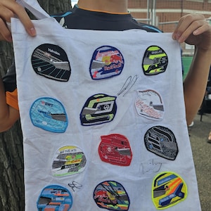 Puede incluir: Bolso de mano blanco con parches bordados de cascos de carreras en varios colores y diseños. Los parches incluyen logotipos y texto. El bolso lo sostiene una persona.