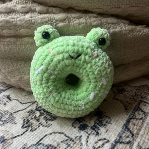 May include: A green crocheted donut shaped plush toy with white frosting and black eyes. The toy is shaped like a frog with a small hole in the centre.
