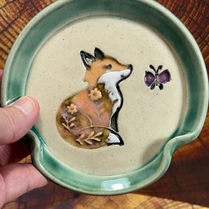 May include: A round ceramic spoon rest with a speckled cream center and a sage green rim. The center features a raised, hand-painted orange and white fox with floral details on its body, positioned next to a small purple butterfly.