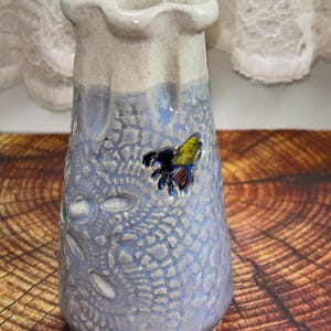 May include: A ceramic vase with a ruffled rim, featuring a speckled cream-colored top and a textured light blue base. The body is decorated with an embossed honeycomb pattern and a small, raised, hand-painted bee with yellow and black wings.