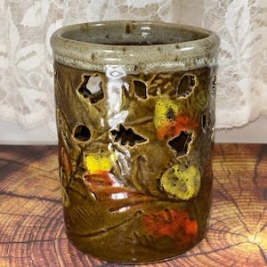 May include: A cylindrical ceramic vessel with a glossy brown glaze, featuring cut-out leaf shapes and raised, textured leaf motifs in shades of yellow, orange, and red. The rim has a lighter, speckled beige glaze, and the body has a smooth finish.