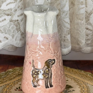 May include: A ceramic vase with a light pink and white color scheme. The vase features a dog illustration on the front. The top of the vase has a ruffled edge.