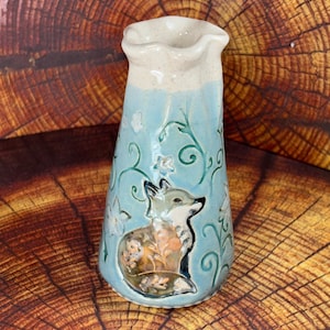 May include: A ceramic vase with a flared, wavy rim, featuring a light blue body decorated with embossed, swirling vine patterns and small flowers. A detailed, raised fox figure with orange and brown tones is centered on the front of the vase.