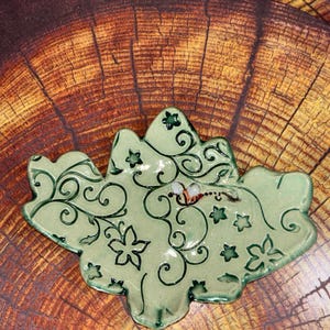 May include: A light green ceramic dish shaped like a leaf with dark green swirling vine patterns and small star cutouts. A small, painted bee with white wings and a brown body is positioned on the surface of the dish, adding detail to the design.