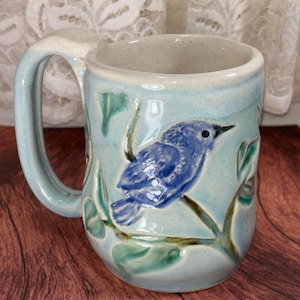 May include: A ceramic mug with a light blue glaze featuring a raised, hand-painted dark blue bird perched on a branch with green leaves. The mug has a thick, curved handle and a speckled off-white interior, showcasing a textured finish.
