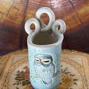 May include: A light blue ceramic vessel featuring a raised relief of an owl perched on a branch. The top of the container has three decorative looped handles. The surface has a speckled finish with embossed scrollwork patterns surrounding the owl.