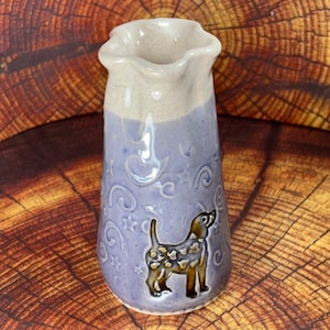 May include: A ceramic pitcher with a wavy rim, featuring a two-tone glaze of cream at the top and lavender on the body. The lavender section is decorated with embossed stars and swirls, and a brown dog figure is centered on the front surface.