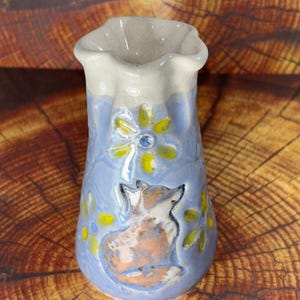 May include: A small, ceramic vase with a light blue body, featuring a fox and floral design. The vase has a ruffled top and a speckled, off-white rim. Yellow and blue flowers are painted around the fox. The vase is approximately 4 inches tall.