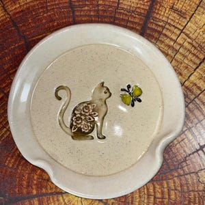 May include: A round, cream-colored ceramic spoon rest with a speckled finish. The center features a raised, brown-toned cat silhouette decorated with a white floral pattern, positioned next to a small, yellow and black butterfly accent.