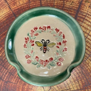 May include: A round ceramic spoon rest with a glossy green rim and a cream-colored center. The center features a hand-painted bee surrounded by a circular wreath of small red and green floral details, creating a decorative kitchen accessory.