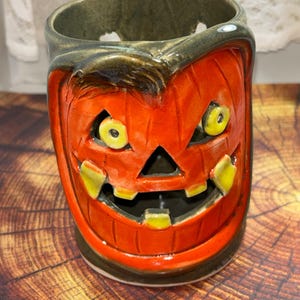 May include: A cylindrical ceramic jack-o'-lantern candle holder with a glossy orange finish, featuring a carved face with yellow eyes and teeth, a dark green rim, and cutouts for the eyes, nose, and mouth to allow light to pass through the vessel.