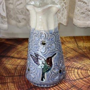 May include: A ceramic pitcher with a flared, speckled cream-colored rim and a periwinkle blue body. The exterior features a raised, embossed floral pattern and a central relief of a hummingbird with green, brown, and white plumage details.