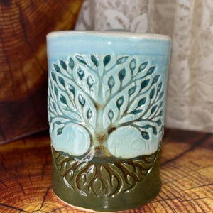 May include: A ceramic mug with a tree of life design. The mug features a light blue top, a teal tree design, and a dark green base. The tree's branches and roots are intricately carved into the mug's surface, creating a textured appearance.