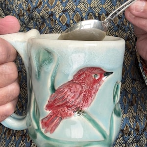 May include: A ceramic mug with a light blue and green glaze featuring a raised, textured red bird perched on a branch. The mug has a sturdy handle and contains a silver spoon with the inscription Towle Sterling visible on the handle.