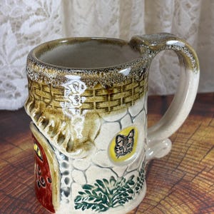 May include: A ceramic mug shaped like a whimsical cottage. The mug features a red door, a yellow window, and a cat illustration. The exterior is textured with stone and brick details, and the handle is a curved design.