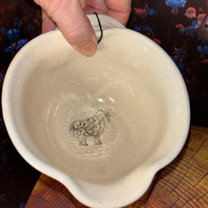 May include: Cream-colored ceramic bowl with a spout and a small, decorative sheep design inside. The bowl has a speckled texture and a black string attached for hanging. The bowl is held by a hand.