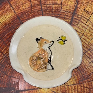 May include: A ceramic spoon rest with a cream-colored base and a raised design of a fox with floral accents and a yellow butterfly. The spoon rest has a white rim and two small handles.