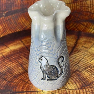 May include: A ceramic vase with a flared, wavy rim and a speckled cream-colored top that transitions into a light blue, textured body. The surface features a raised, dark-toned cat silhouette with a swirling pattern on its side and a long, curved tail.