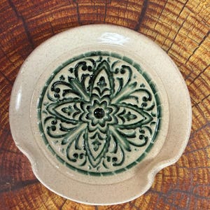 May include: A ceramic spoon rest with a speckled beige finish and a central embossed design in dark green. The design features a floral pattern. The spoon rest has a curved shape with a small lip on one side.