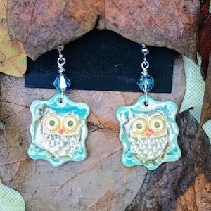 May include: A pair of handmade owl earrings. Each earring features a square, turquoise-edged pendant with a white owl design. The owl has yellow eyes and a red beak. The earrings hang from silver-colored hooks with blue crystal accents.