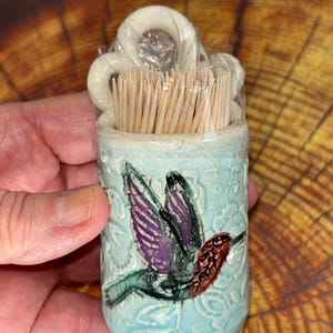 May include: A small, light blue ceramic toothpick holder with a hummingbird design. The hummingbird has purple wings and a red body. The holder is filled with wooden toothpicks and has a decorative floral pattern.