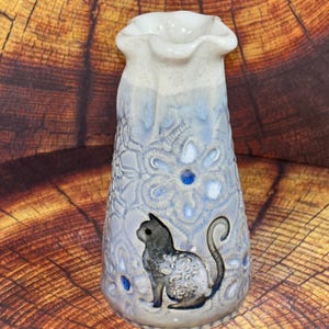 May include: A ceramic vase with a ruffled rim, featuring a light blue and white glaze with an embossed floral pattern. A dark gray cat silhouette is centered on the front, accented with small, deep blue circular inlays throughout the design.
