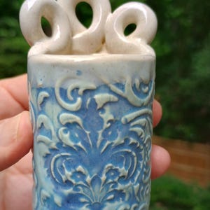 May include: A ceramic vase with a blue and white floral design. The vase has a cylindrical shape with a textured surface and three loop-shaped handles at the top. The color palette includes shades of blue and white.