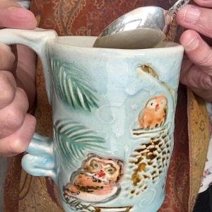May include: A light blue ceramic mug featuring embossed details of two owls perched on branches with pinecones and pine needles. The mug has a sculpted handle and a glossy finish, with the owls painted in shades of brown, orange, and red.