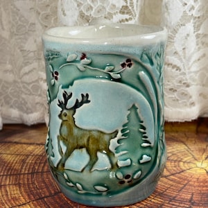 May include: A ceramic mug with a glossy light blue and green glaze featuring a raised relief design of a brown deer standing in a forest with pine trees and small red berries. The mug has a smooth, rounded rim and a textured, sculpted surface.