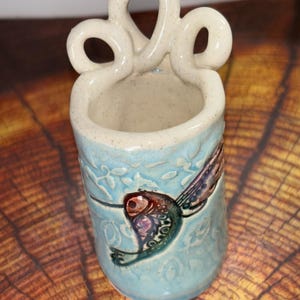 May include: A ceramic vase with a light blue exterior and a hummingbird design. The vase has a textured surface and a decorative handle. The hummingbird is depicted with red, purple, and green details. The vase is approximately 5 inches tall.