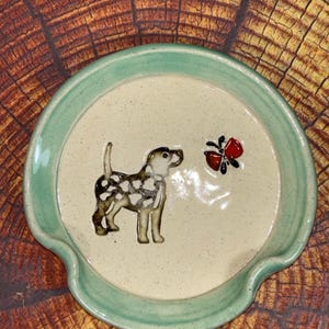 May include: A round ceramic spoon rest with a speckled cream-colored center and a sage green rim. The center features a raised, hand-painted illustration of a brown and white dog standing and looking toward a small, bright red butterfly with black wings.