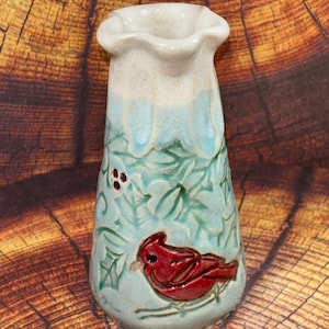 May include: A ceramic vase with a flared, wavy rim featuring a glossy white top section and a light blue base. The body is decorated with embossed green holly leaves and a prominent, raised red cardinal bird perched on a branch.