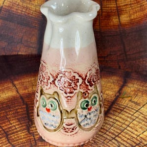 May include: A ceramic vase with a ruffled rim, featuring a cream-colored top and a light pink base. The body is decorated with embossed lace patterns and two owl figures with green eyes, red beaks, and blue and white patterned bodies.