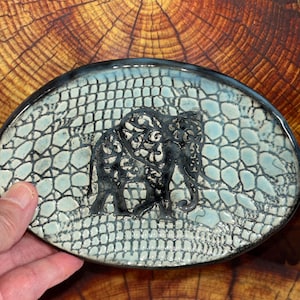 May include: An oval ceramic dish featuring a black elephant silhouette with intricate cut-out patterns. The surface has a light blue, crackled glaze texture resembling reptile skin, framed by a dark, smooth, and glossy black rim.